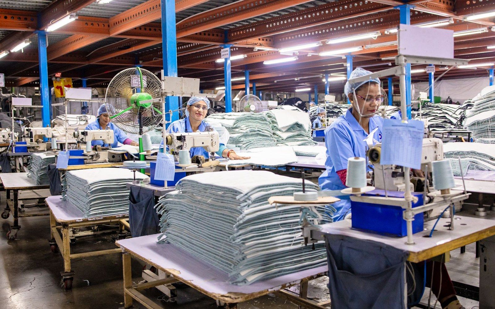 Adult machinists at rows of industrial sewing machines on a garment factory floor, overhead fluorescent light, bolts of fabric stacked at the end of each row