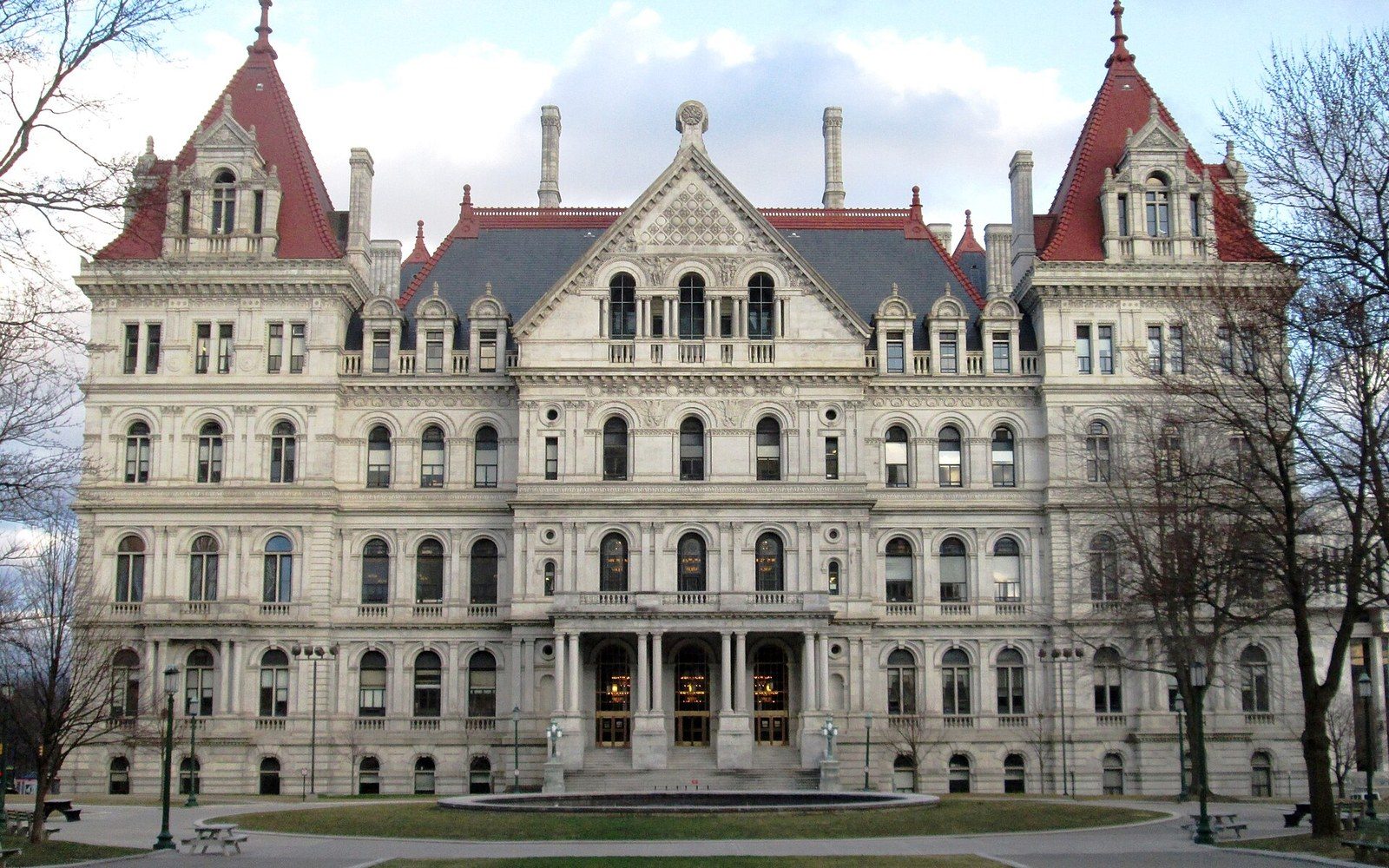 Grand stone capitol building with red-roofed towers and arched windows facing a lawn