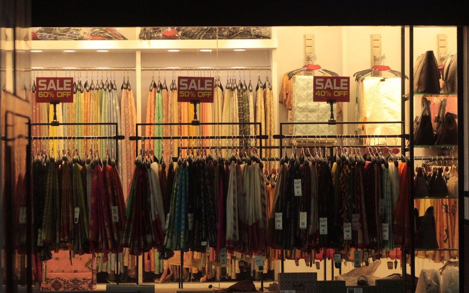 A European high-street shop window showing a price display and rail of hanging garments, adult shoppers reflected in the glass