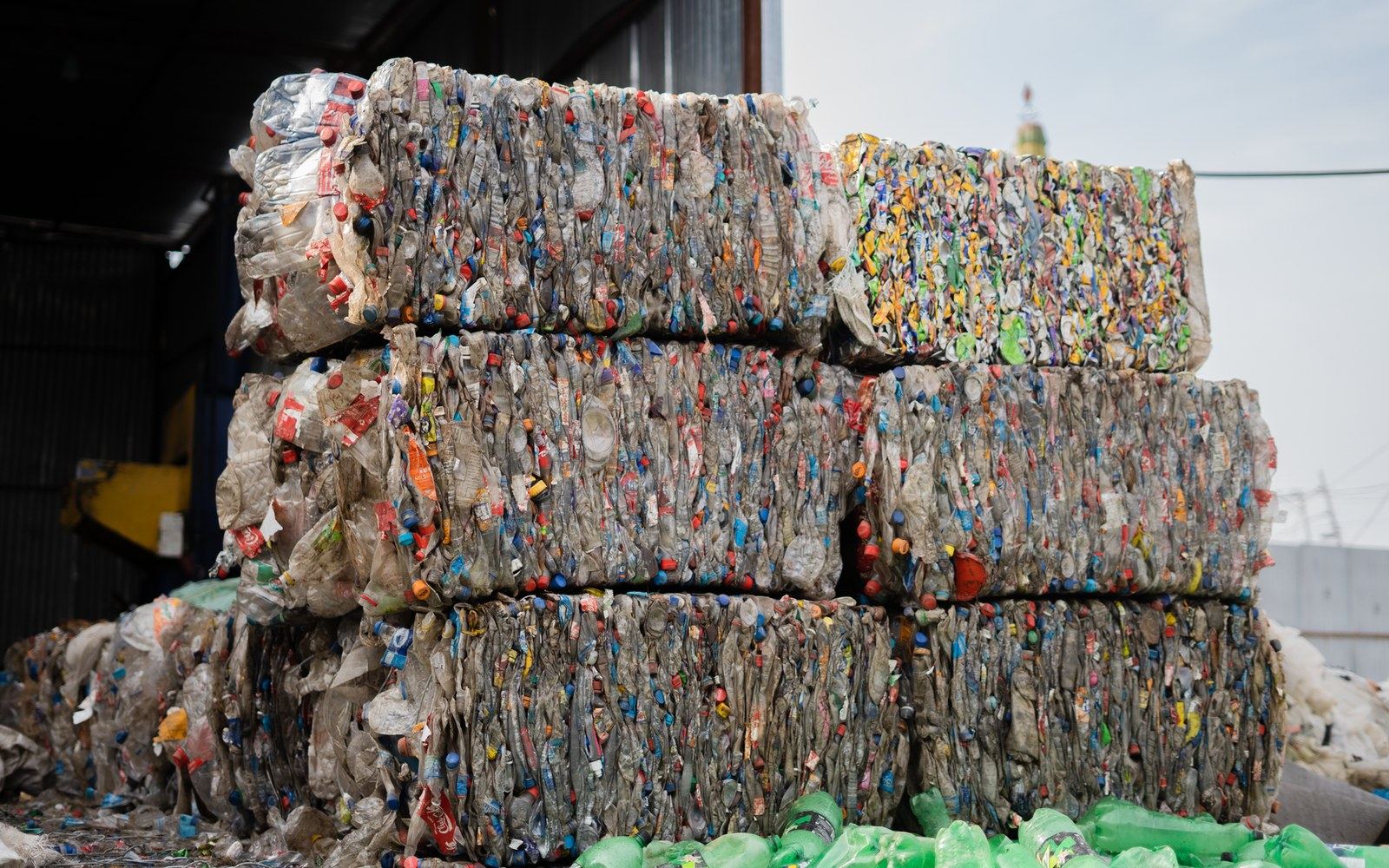 Compressed bales of mixed textiles in a recycling facility yard, forklift visible at the edge of frame, daylight overhead