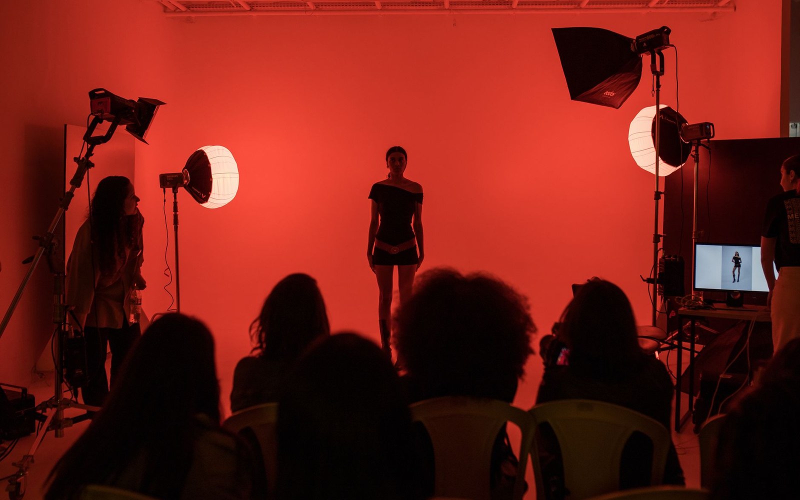 A fashion runway photographed from the press position at working distance, adult model mid-stride, seated audience and photographers visible on both sides